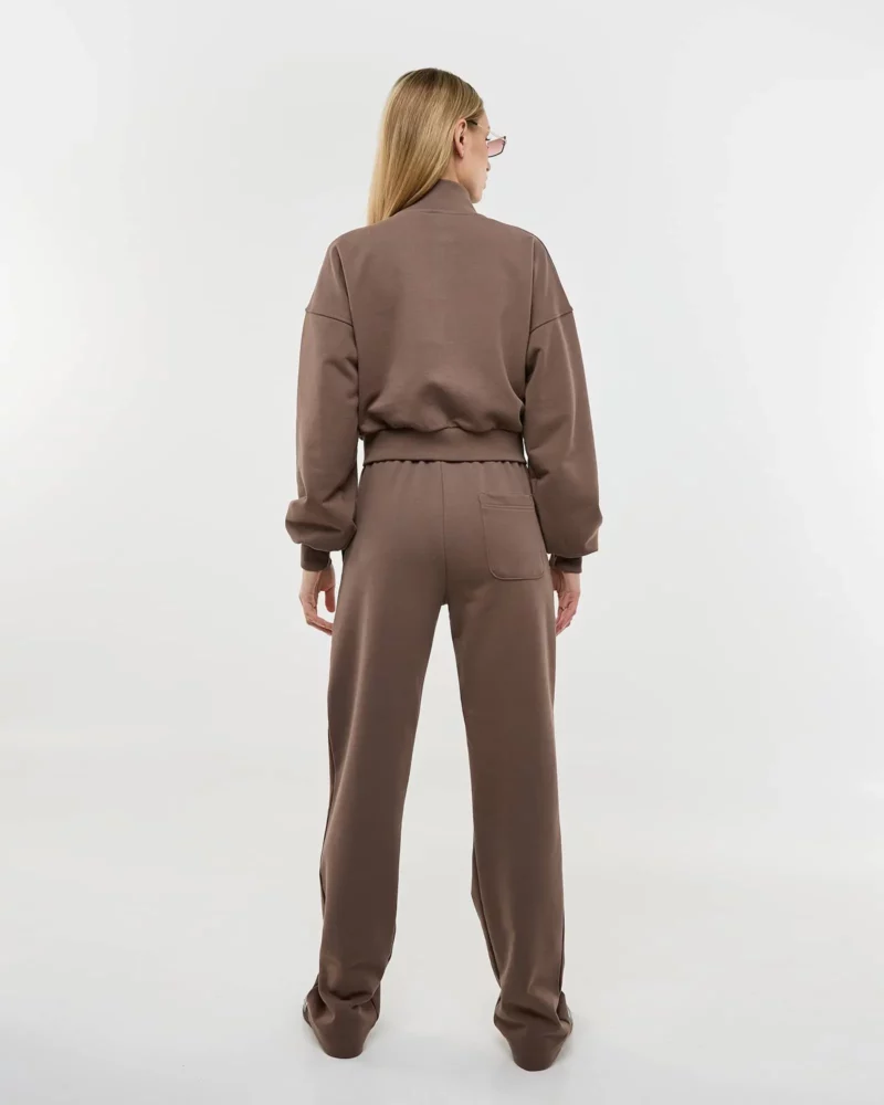 Šviesiaplaukė mergina dėvi rudą laisvalaikio kostiumėlį su užtrauktuku ir stovi atsukusi nugarą