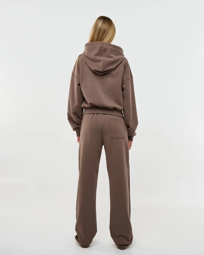 Mergina stovi atsukusi nugarą ir vilki rudos spalvos laisvalaikio kostiumą su tiesiomis kelnėmis ir džemperiu su užtrauktuku